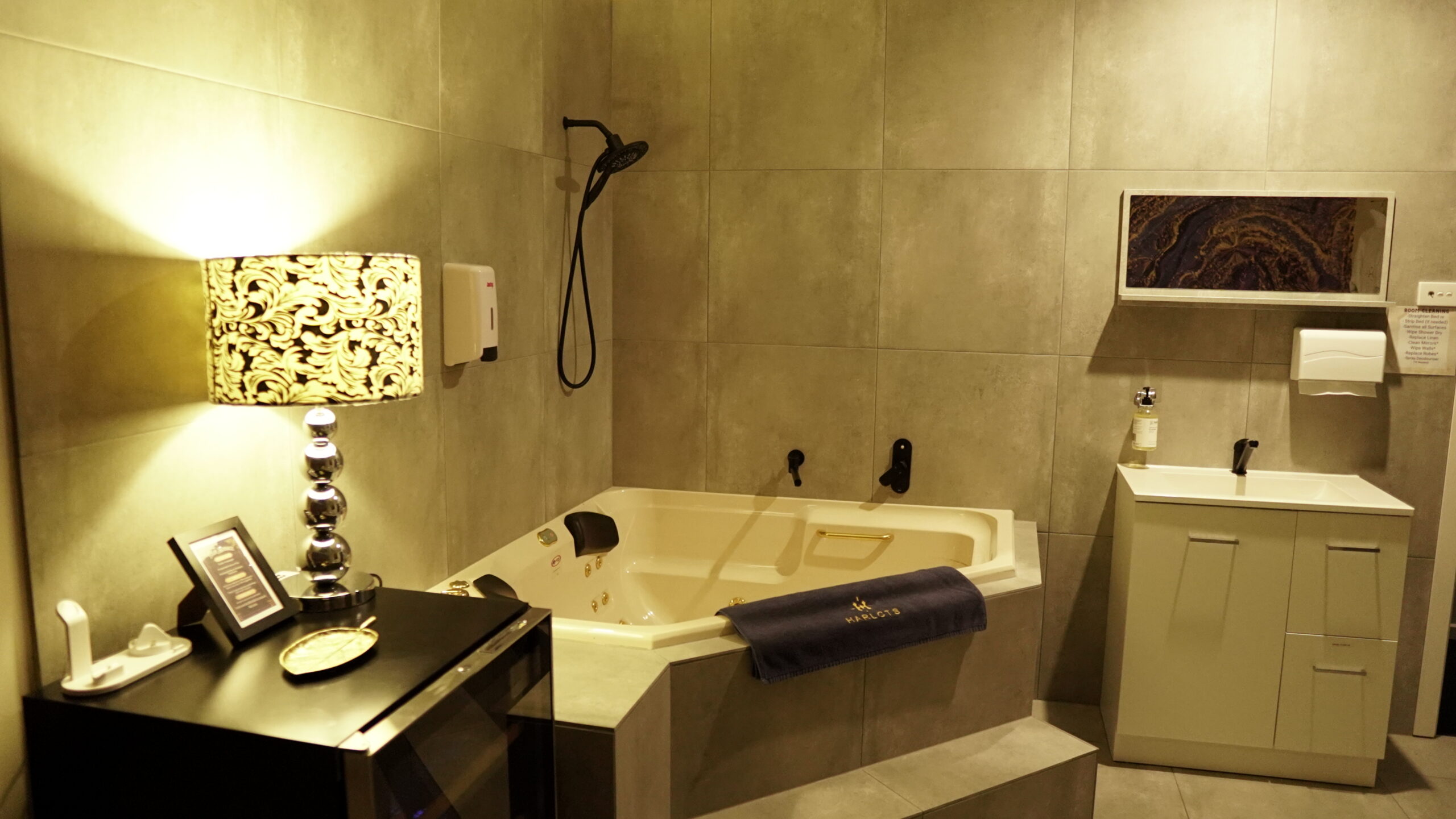 visiting a Harlots brothel Modern private bathroom suite featuring a built-in spa bath, sleek vanity, wall shower, and navy towel embroidered with the Harlots logo